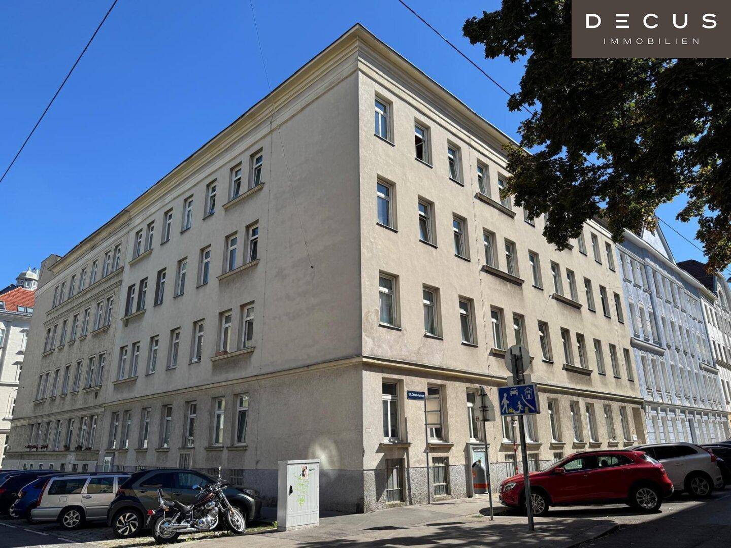 Historisches Eckgebäude mit vielen Fenstern und Bäumen im Vordergrund unter blauem Himmel in urbaner Umgebung.