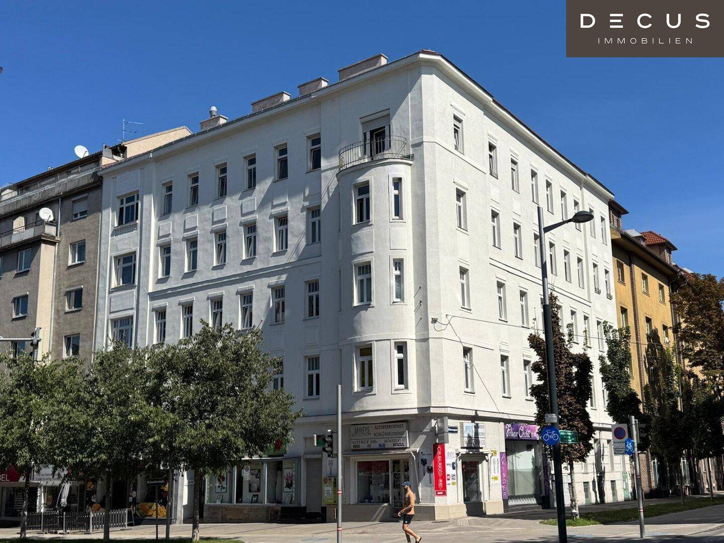 Weißes Eckgebäude mit klassischer Architektur und Geschäften im Erdgeschoss an einer städtischen Straße.