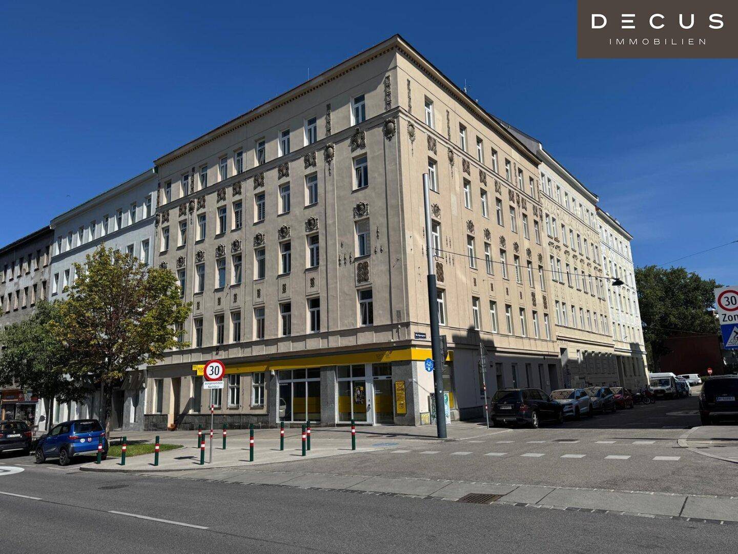 Großes Eckgebäude mit beiger Fassade und einer Postfiliale im Erdgeschoss an einer belebten Kreuzung.