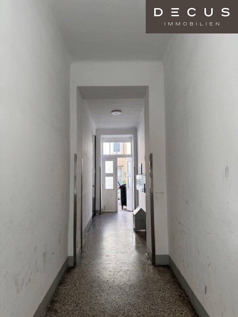 Langer, schmaler Flur mit Terrazzoboden und weiß gestrichenen Wänden, der zu einer Tür führt.