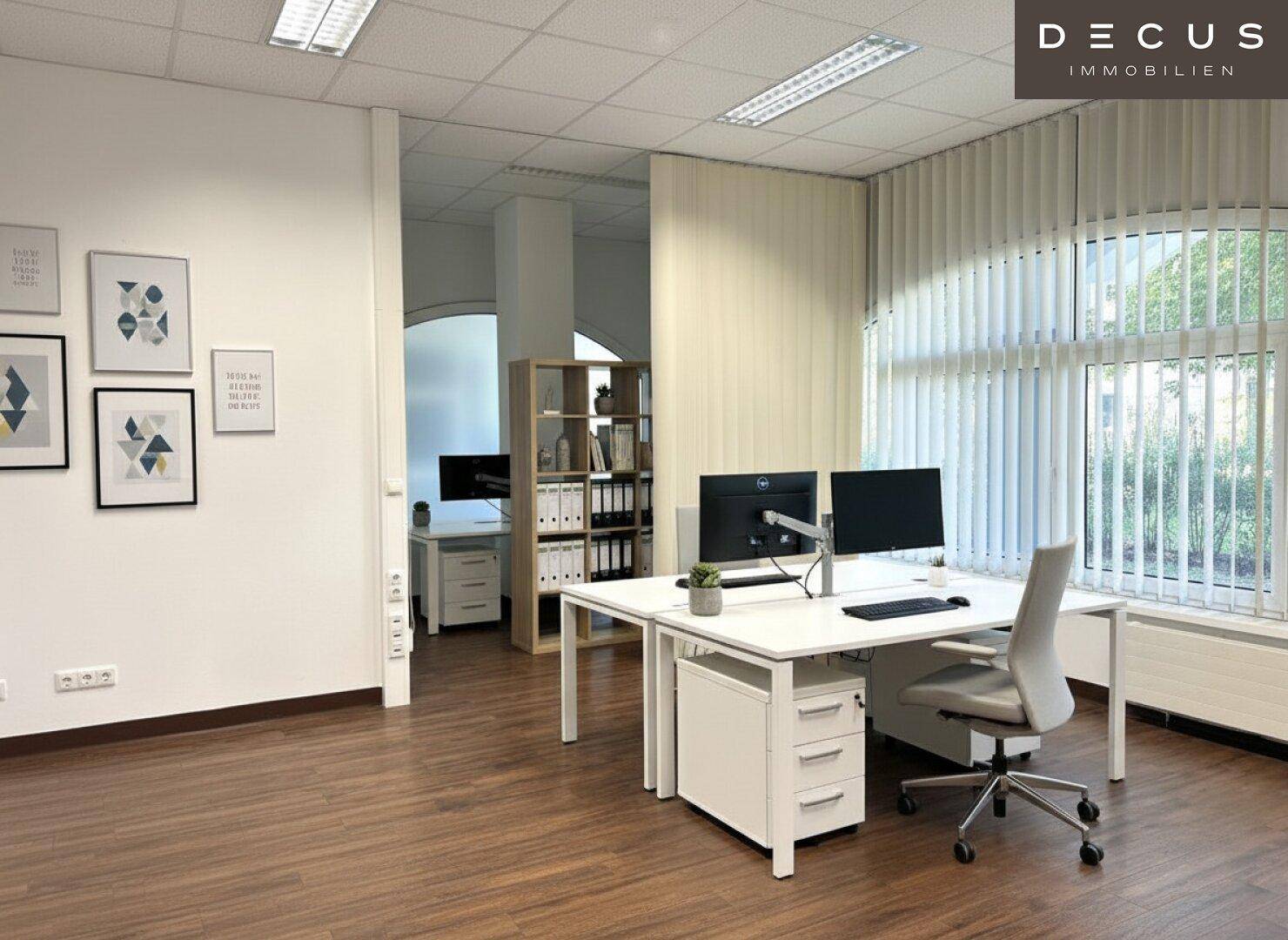 Modern eingerichtetes Büro mit zwei Arbeitsplätzen, Regalen und großen Fenstern mit Jalousien.