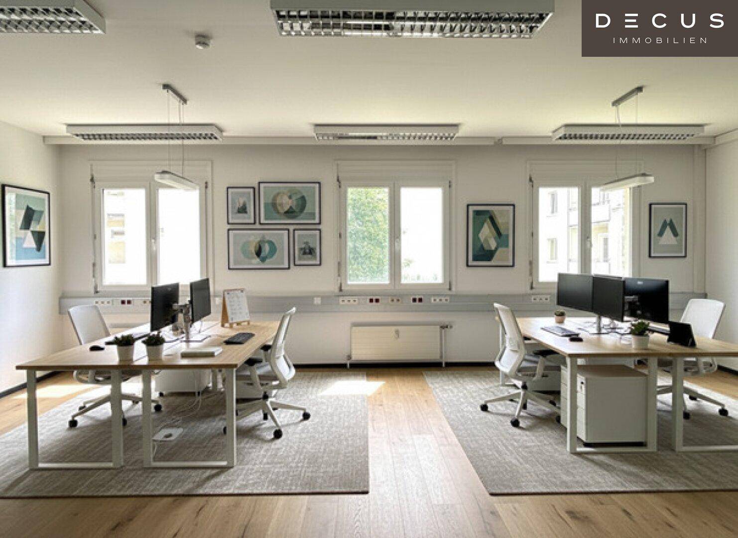 Modernes Büro mit mehreren Arbeitsplätzen, Holzboden und viel Tageslicht durch große Fenster.