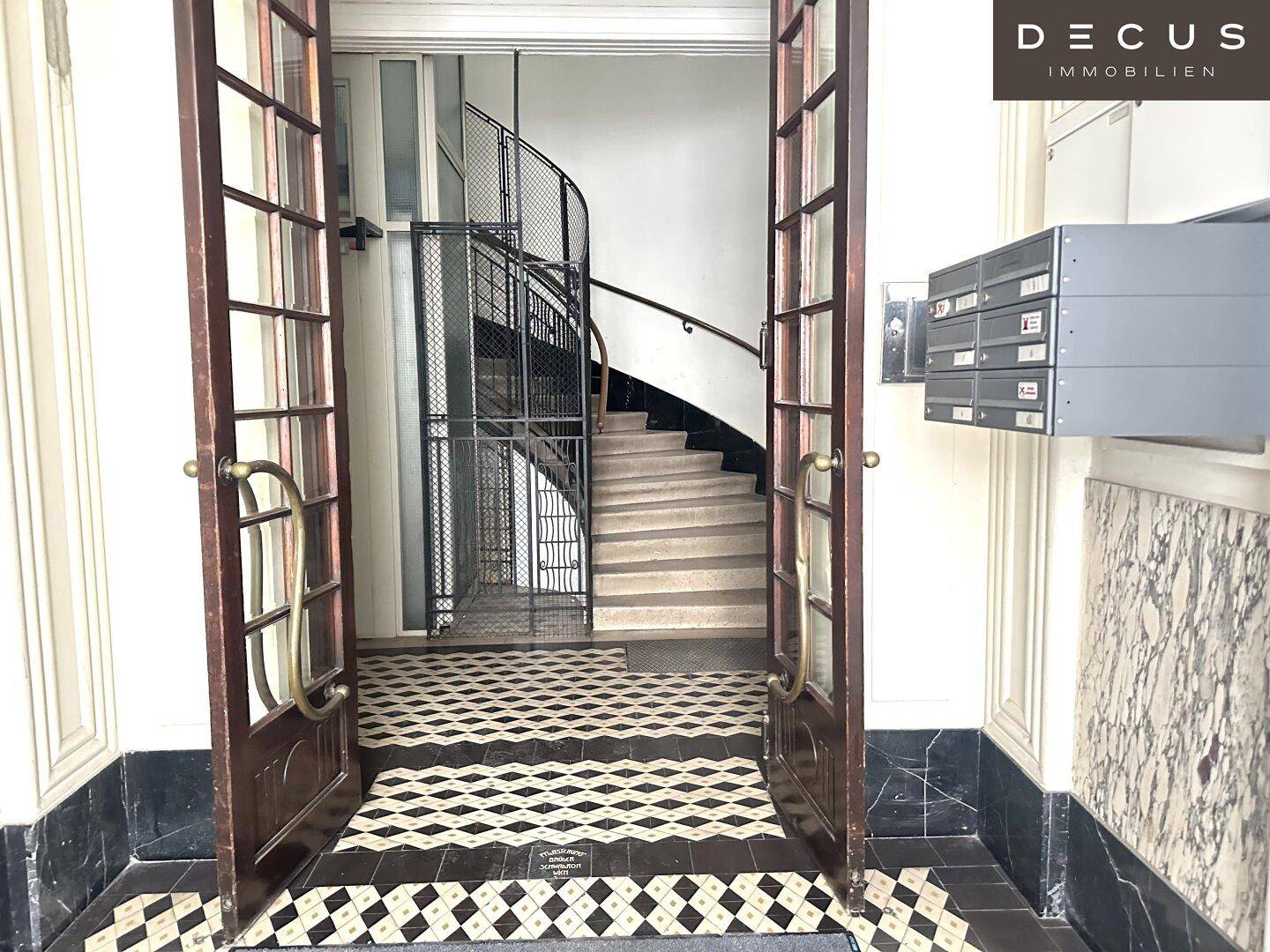 Historischer Eingangsbereich mit gemustertem Fliesenboden, Treppe und altem Gitteraufzug.