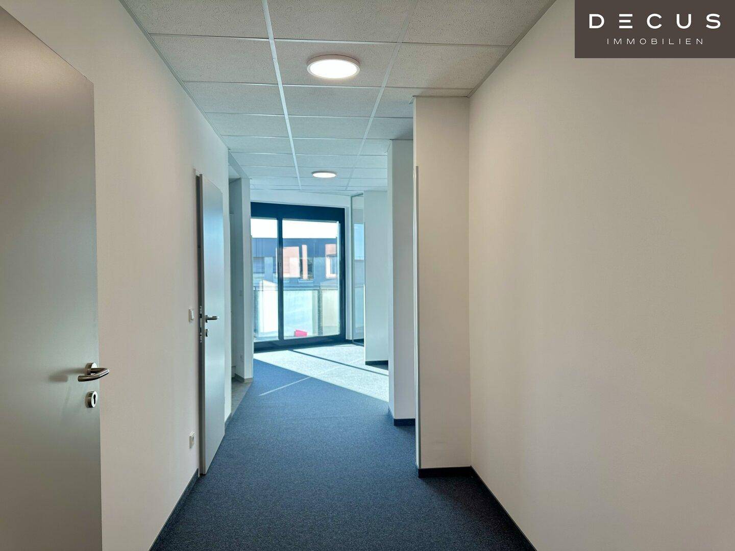 Heller Bürogang mit Teppichboden und Türen zu verschiedenen Räumen, am Ende ein Fenster.