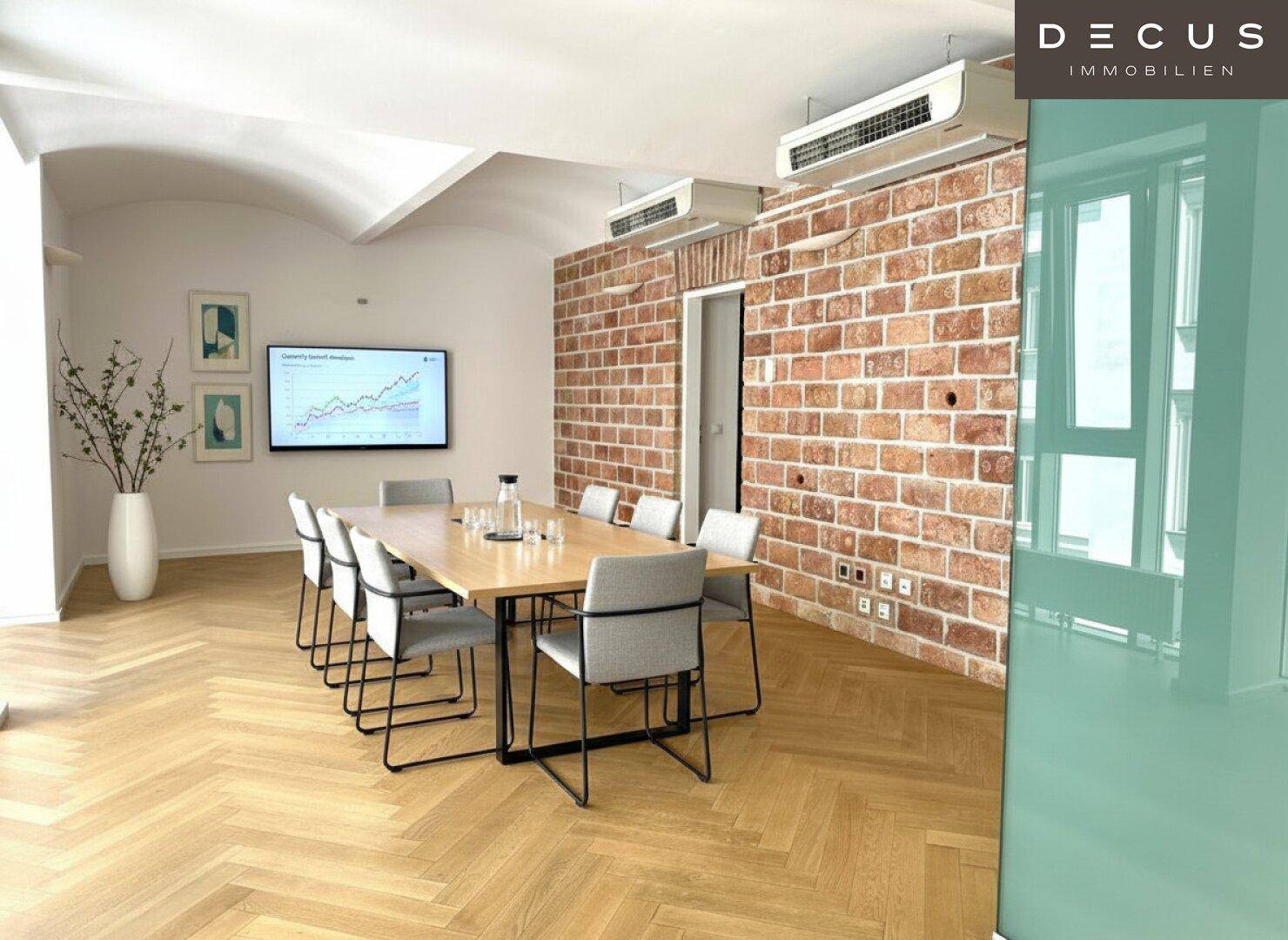 Konferenzraum mit großem Holztisch, Stühlen, Bildschirm und freiliegender Ziegelwand, ideal für Besprechungen.