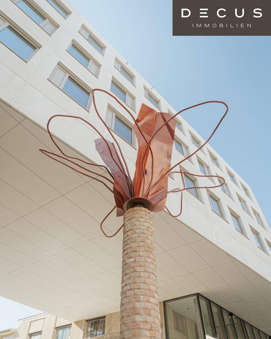 Moderne weiße Gebäudefassade mit einer künstlerischen Metallskulptur davor unter blauem Himmel.