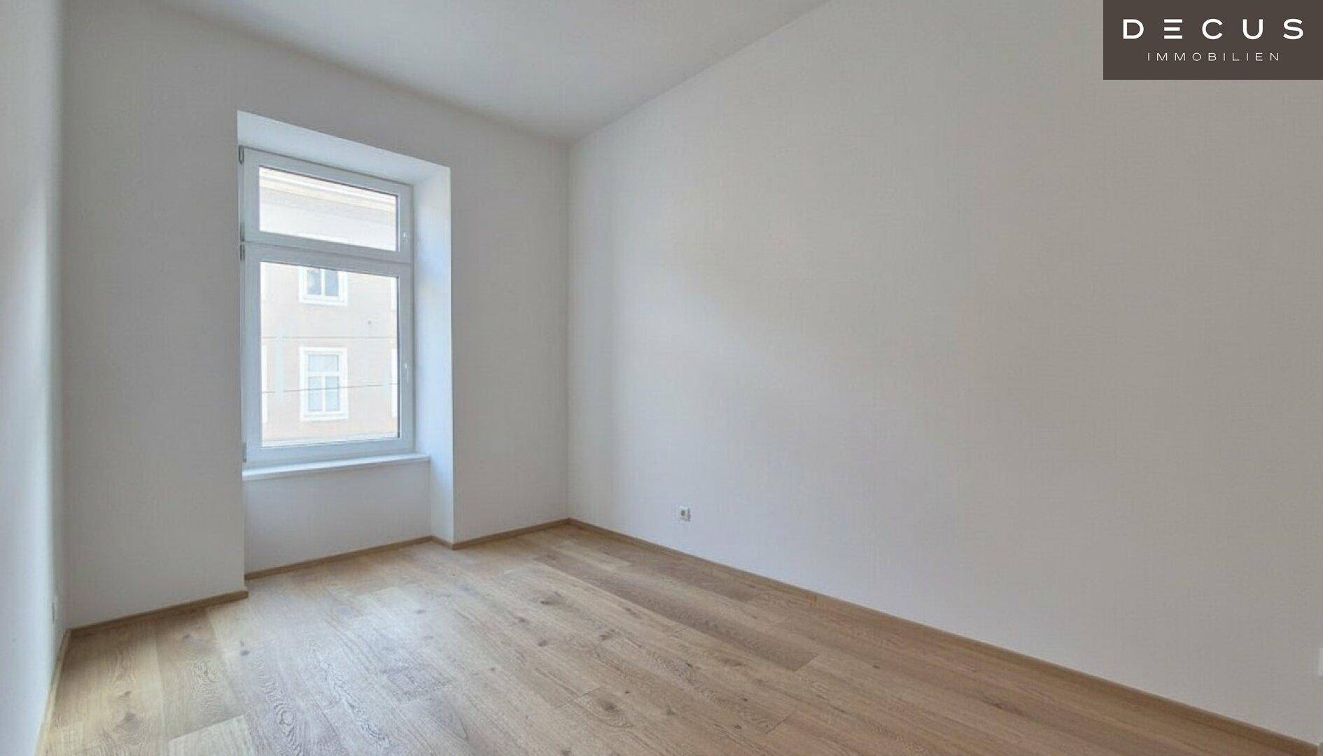 Heller, leerer Raum mit Holzboden und einem Fenster, das viel Tageslicht hereinlässt.