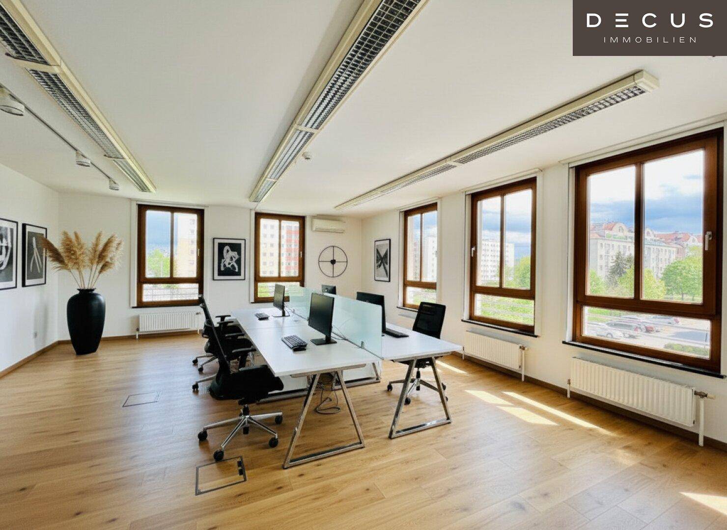 Heller Büroraum mit langer Schreibtischreihe, Bürostühlen und großen Fenstern mit Holzboden.