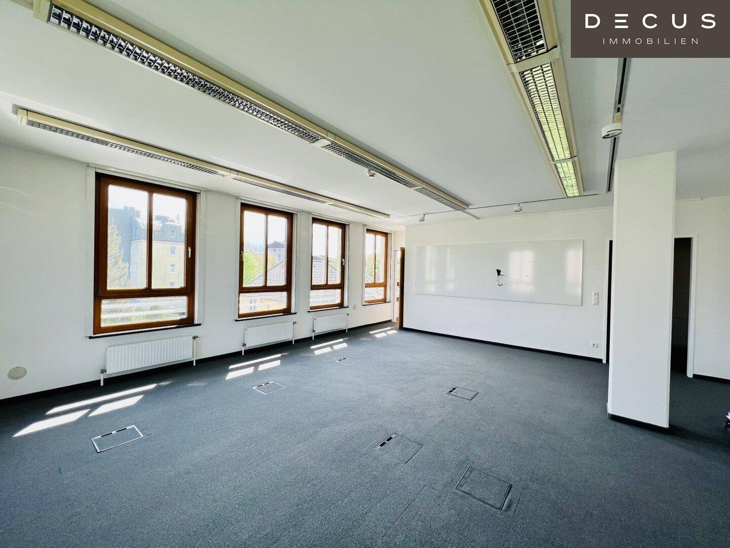 Leerer Büroraum mit dunklem Teppichboden, einer großen Whiteboard-Wand und vielen Fenstern.