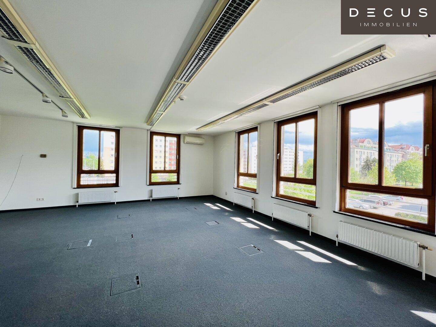 Geräumiger, leerer Büroraum mit dunklem Teppichboden und großen Fenstern, die viel Licht hereinlassen.