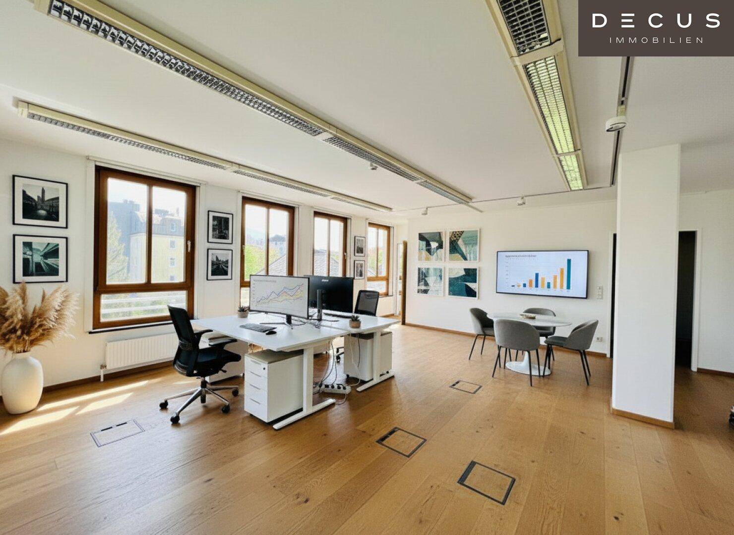 Großes, helles Büro mit Holzboden, mehreren Arbeitsplätzen und Präsentationsbildschirm.