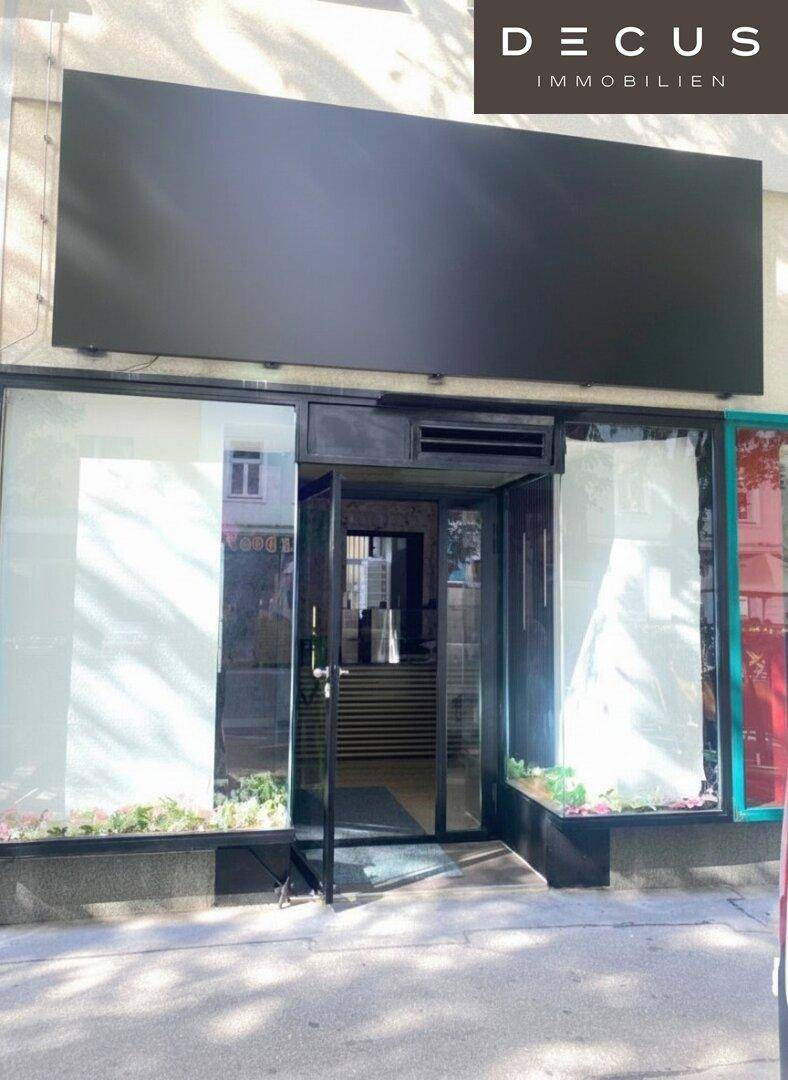 Eingangsbereich eines Geschäfts mit großer Glasfront und schwarzem Schild, einladend gestaltet.