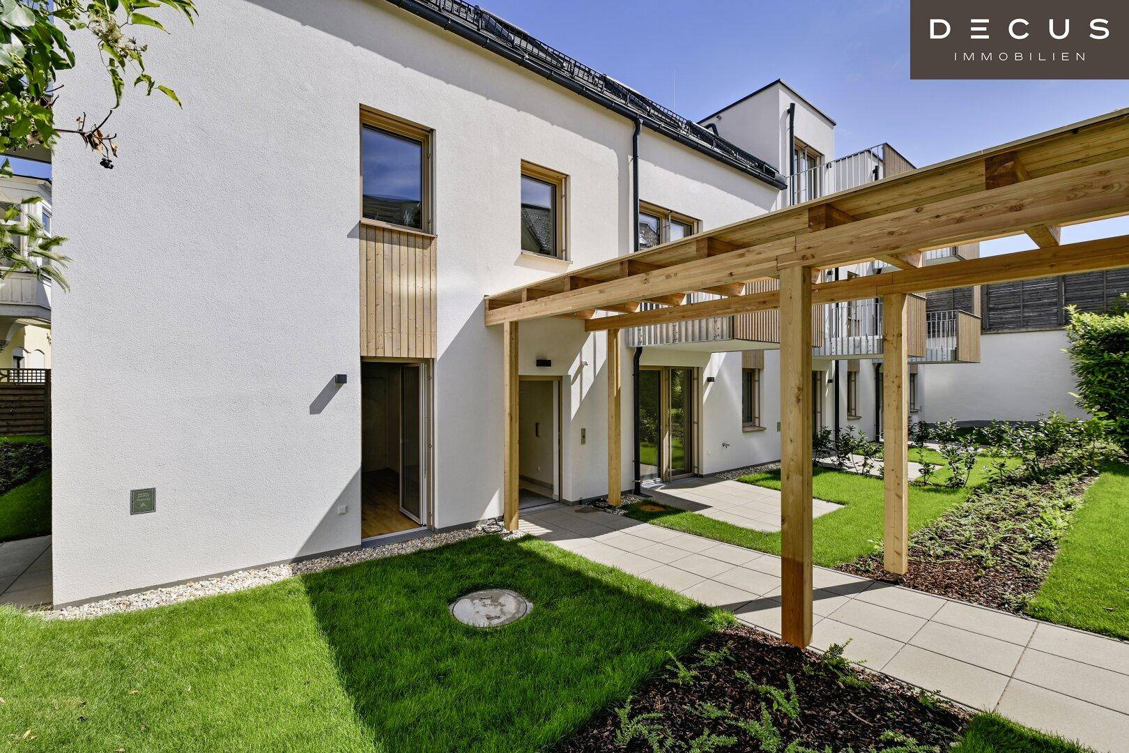 Eingangsbereich der Wohnanlage mit Holzpergola, gepflastertem Weg und grüner Bepflanzung.