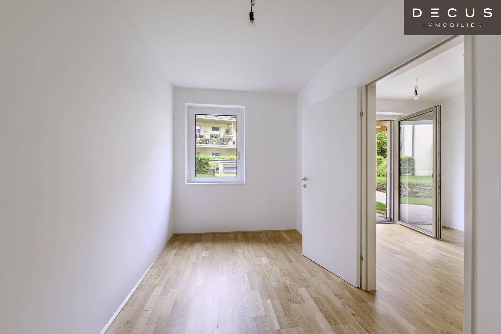 Helles, leeres Zimmer mit Holzboden und Fenster mit Blick ins Grüne.