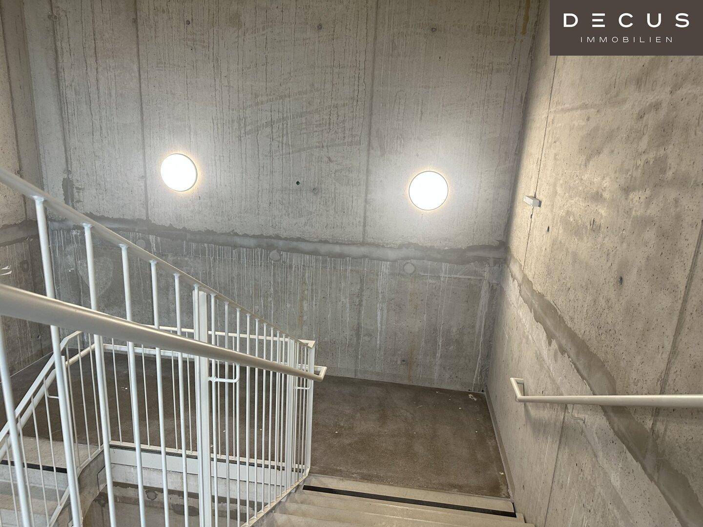 Treppenhaus aus Sichtbeton mit schlichten weißen Geländern und runden Wandleuchten.