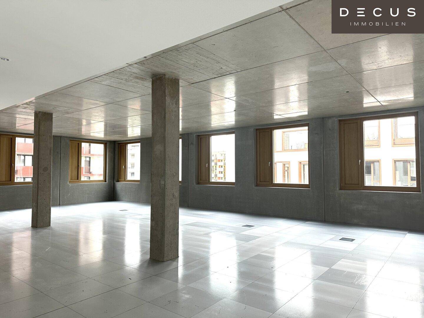 Heller Bürobereich mit vielen Fenstern, Sichtbetonwänden und einem modernen Fliesenboden.