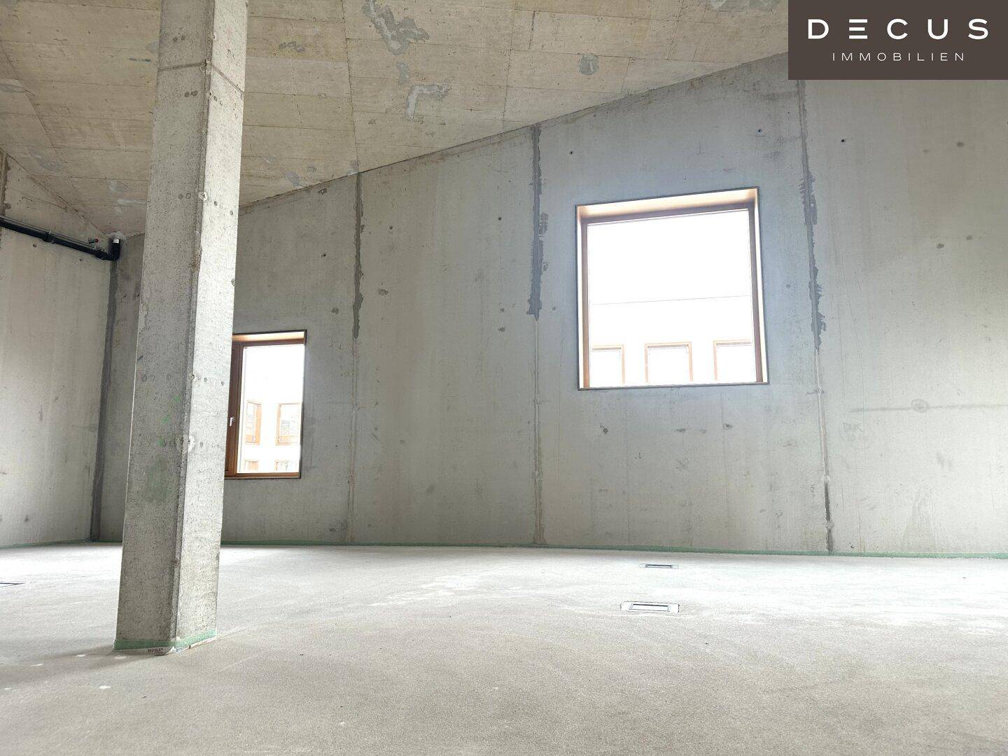 Heller, unmöblierter Raum mit Betonoberflächen, hohen Decken und mehreren Fenstern für Tageslicht.