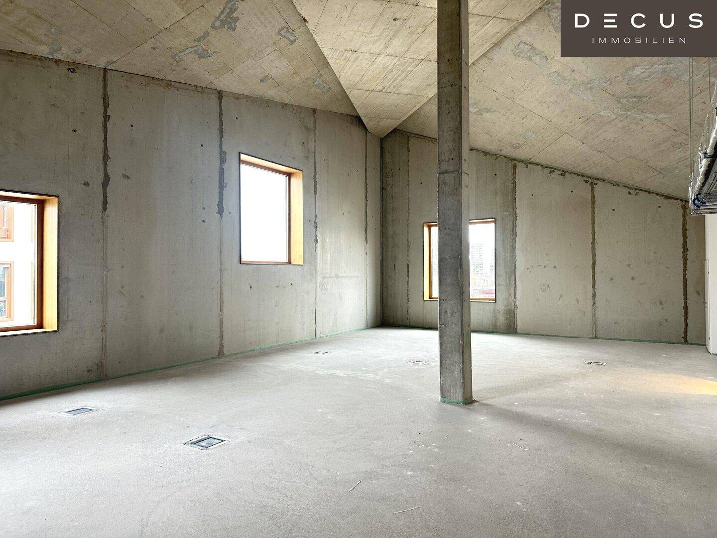 Geräumiger Innenbereich mit schrägen Betondecken, Säulen und mehreren Fenstern, noch im Rohbau.