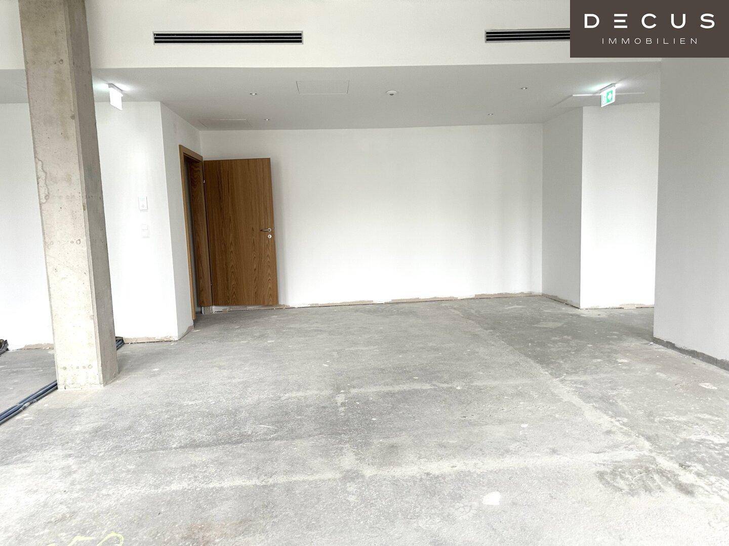 Unmöblierter Innenraum mit Betonboden, weiß gestrichenen Wänden und sichtbaren Lüftungsschlitzen.