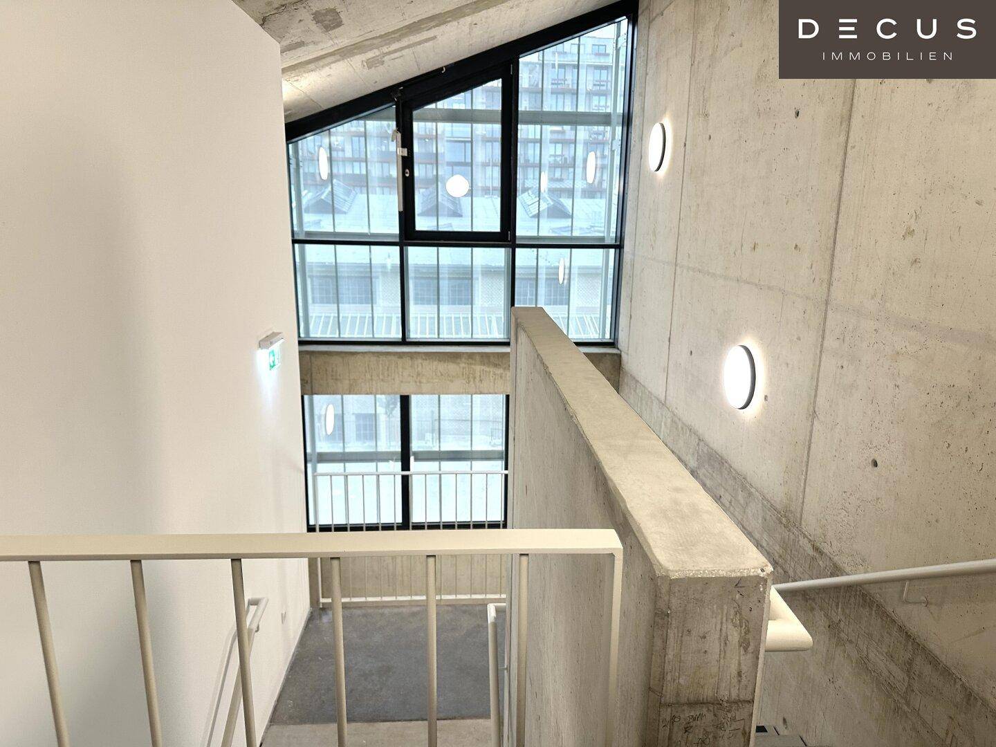 Modernes Treppenhaus mit Betonwänden, Metallgeländer und großen Fenstern, die viel Licht hereinlassen.