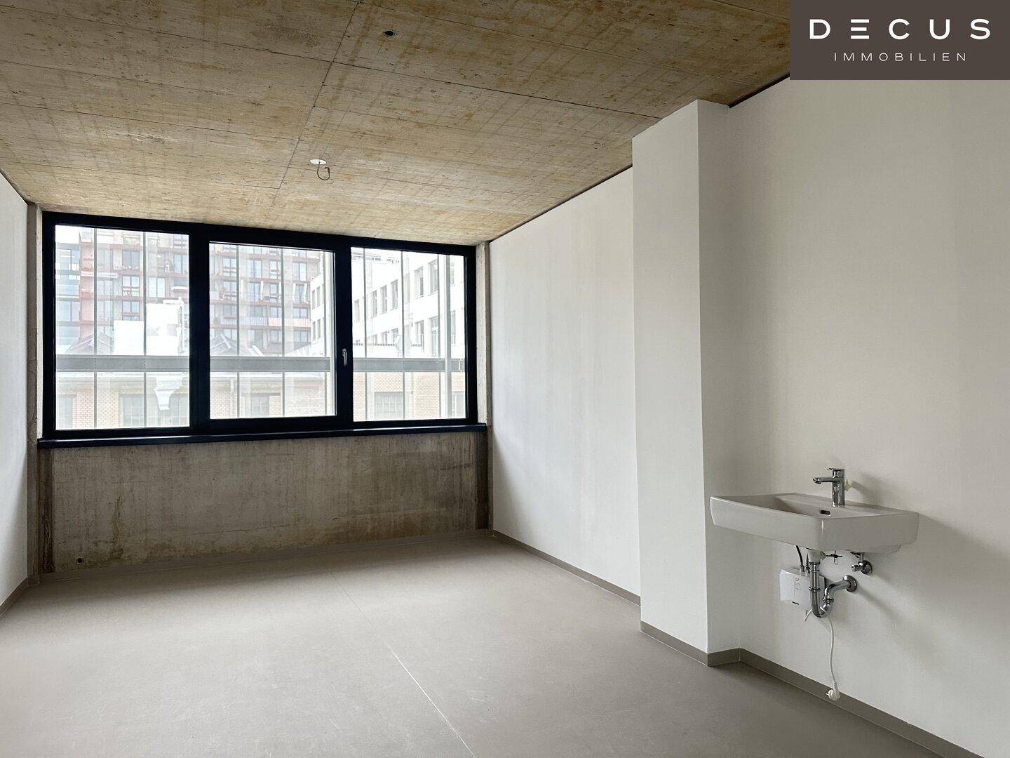 Heller, leerer Raum mit großen Fenstern, Sichtbetondecke und einem Waschbecken an der Wand.
