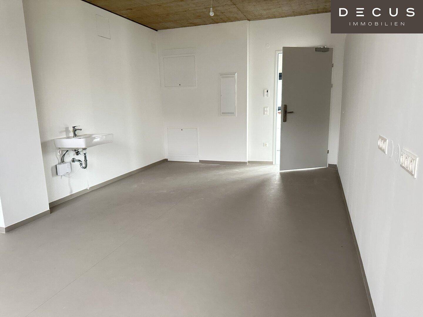 Geräumiger, heller Raum mit Waschbecken, Sichtbetondecke und einer Tür, ideal für Büronutzung.