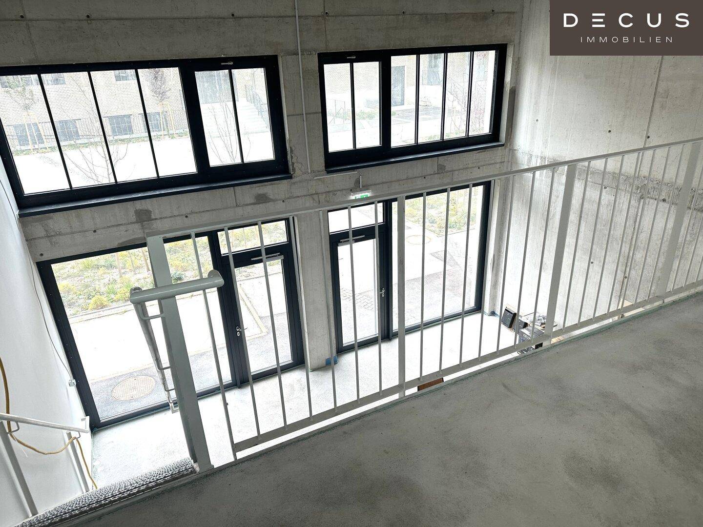 Blick von einer oberen Ebene in einen unfertigen Raum mit hohen Decken und großen Fenstern, die viel Licht spenden.