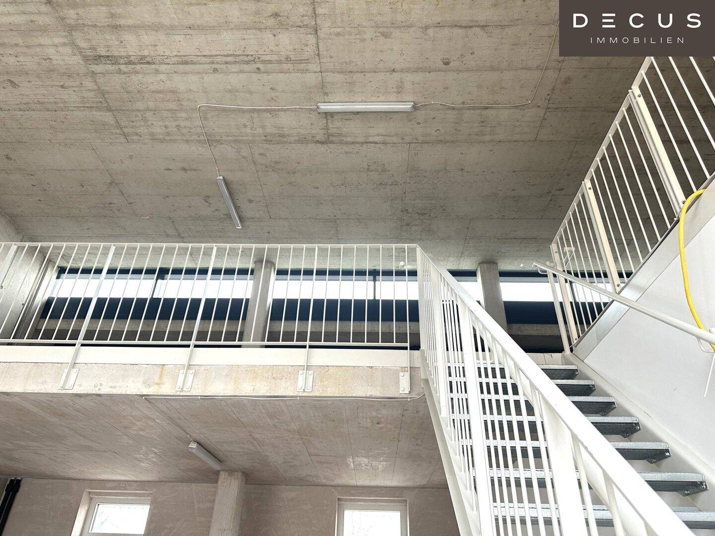 Blick auf eine moderne Metalltreppe und ein Geländer in einem Raum mit hohen Betonwänden und Deckenbeleuchtung.