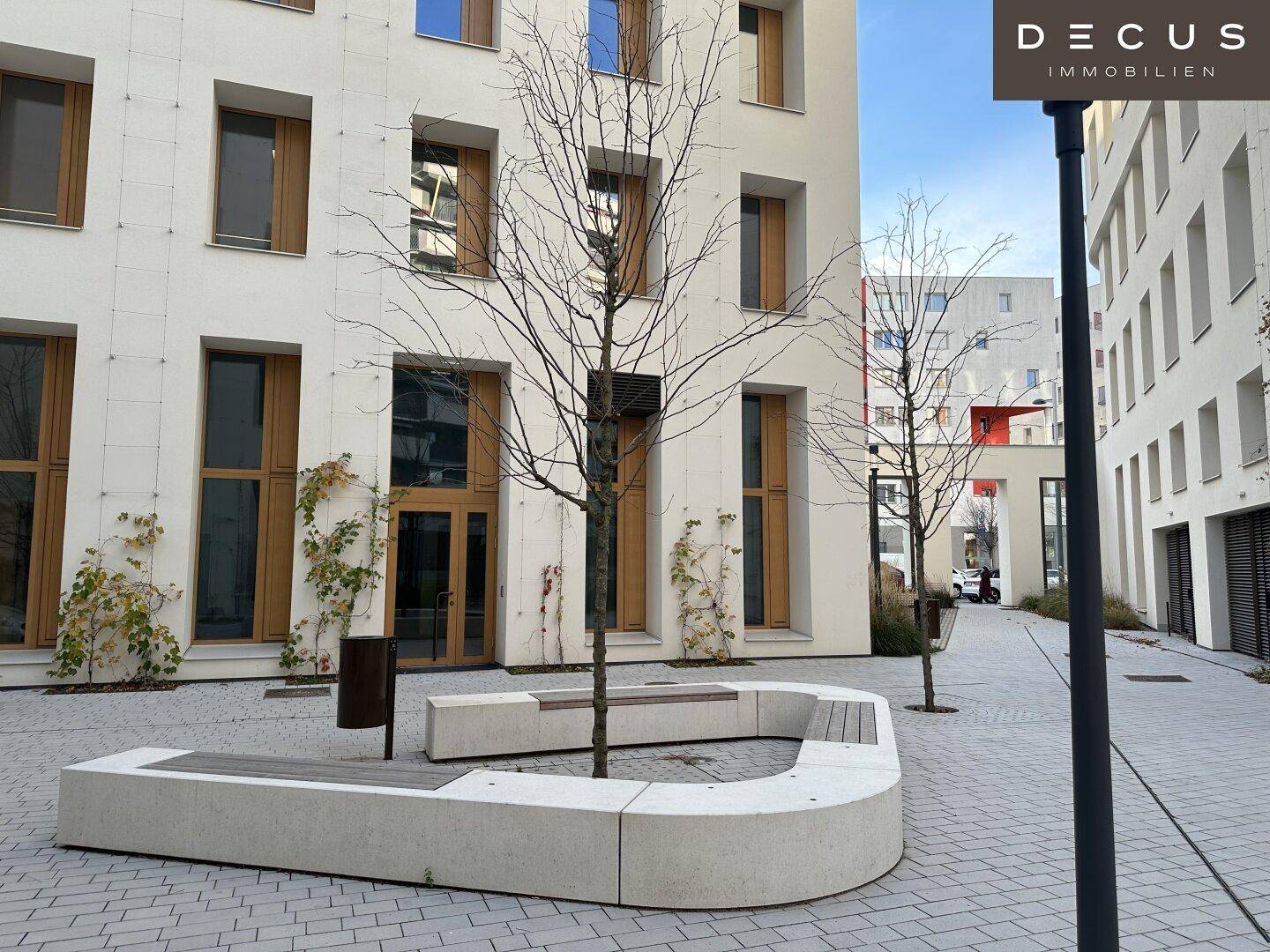 Moderner Gebäudekomplex mit heller Fassade, Bäumen und einer geschwungenen Sitzbank im gepflasterten Innenhof.