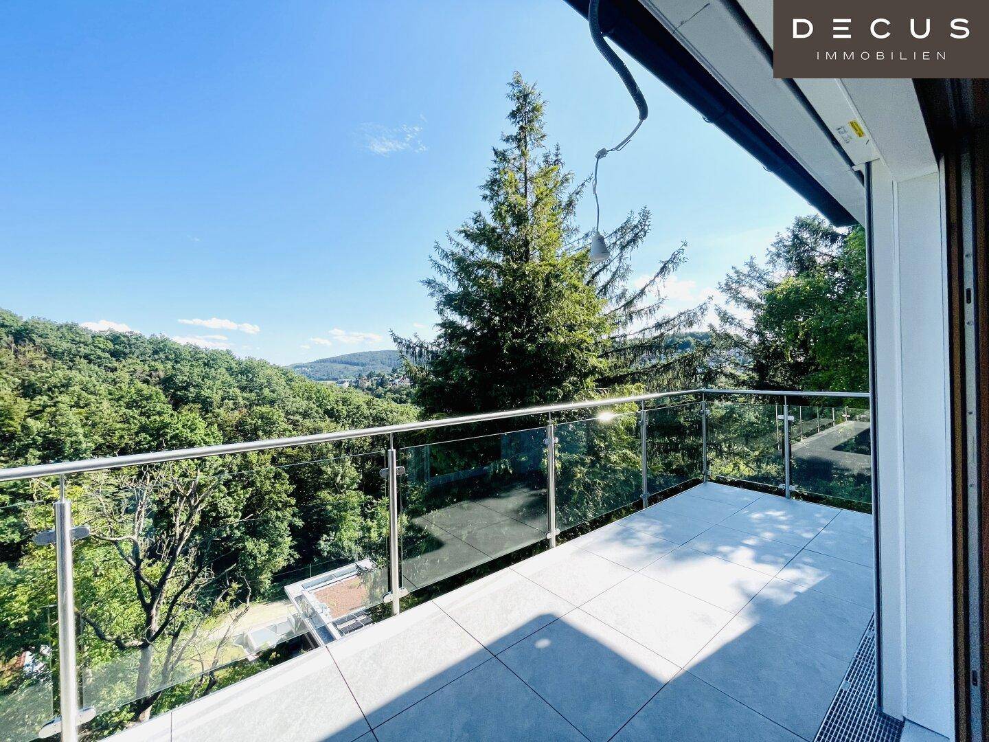 Sonniger Balkon mit Glasgeländer und weitem Blick über die bewaldete Landschaft.