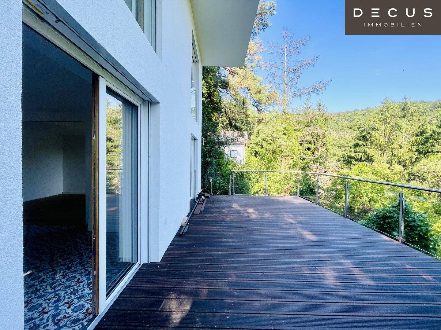 Große Holzterrasse mit Glasgeländer, umgeben von Bäumen und mit Blick ins Grüne.