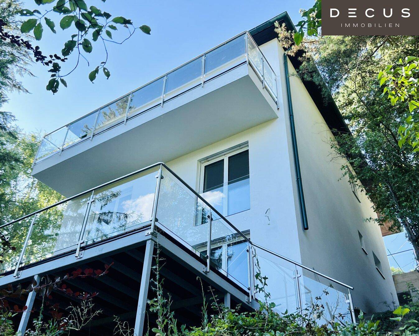 Moderne Hausfassade mit mehreren Balkonen und Glasgeländern, umgeben von Bäumen.