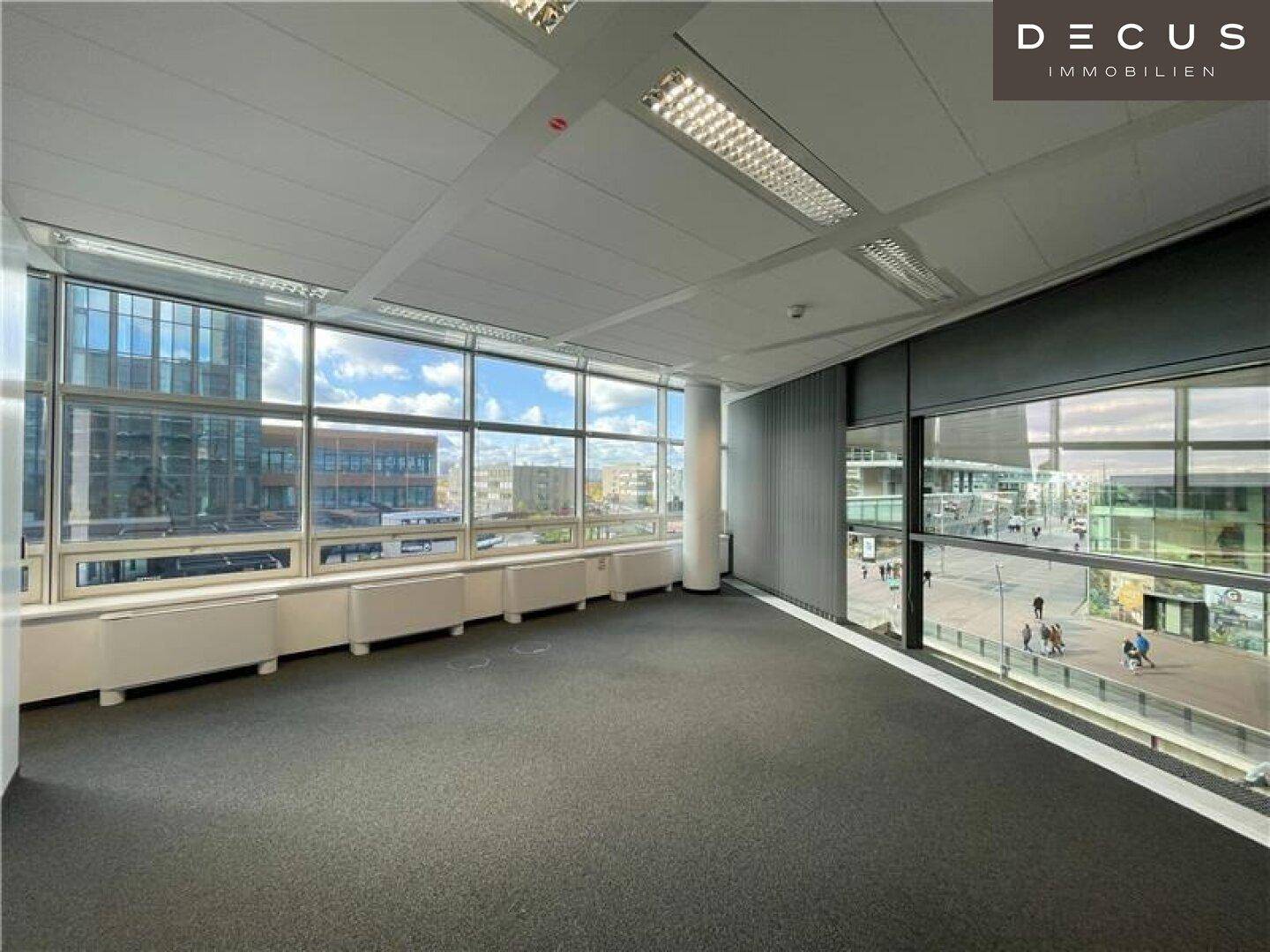 Geräumiges, leeres Büro mit Teppichboden und großen Fenstern, die einen weiten Stadtblick ermöglichen.
