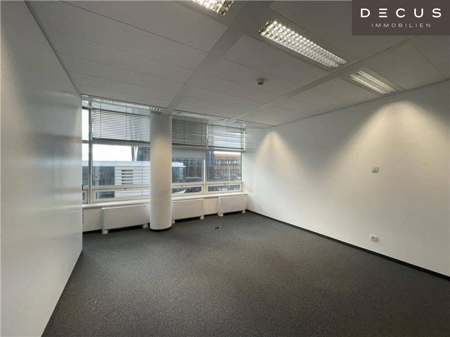 Leeres Büro mit Teppichboden und Fensterfront, die einen urbanen Ausblick bietet.