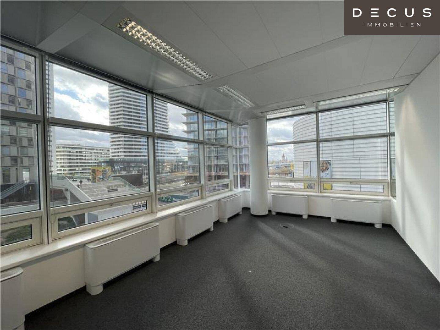 Helles Büro mit Teppichboden und breiter Fensterfront, die einen urbanen Ausblick bietet.