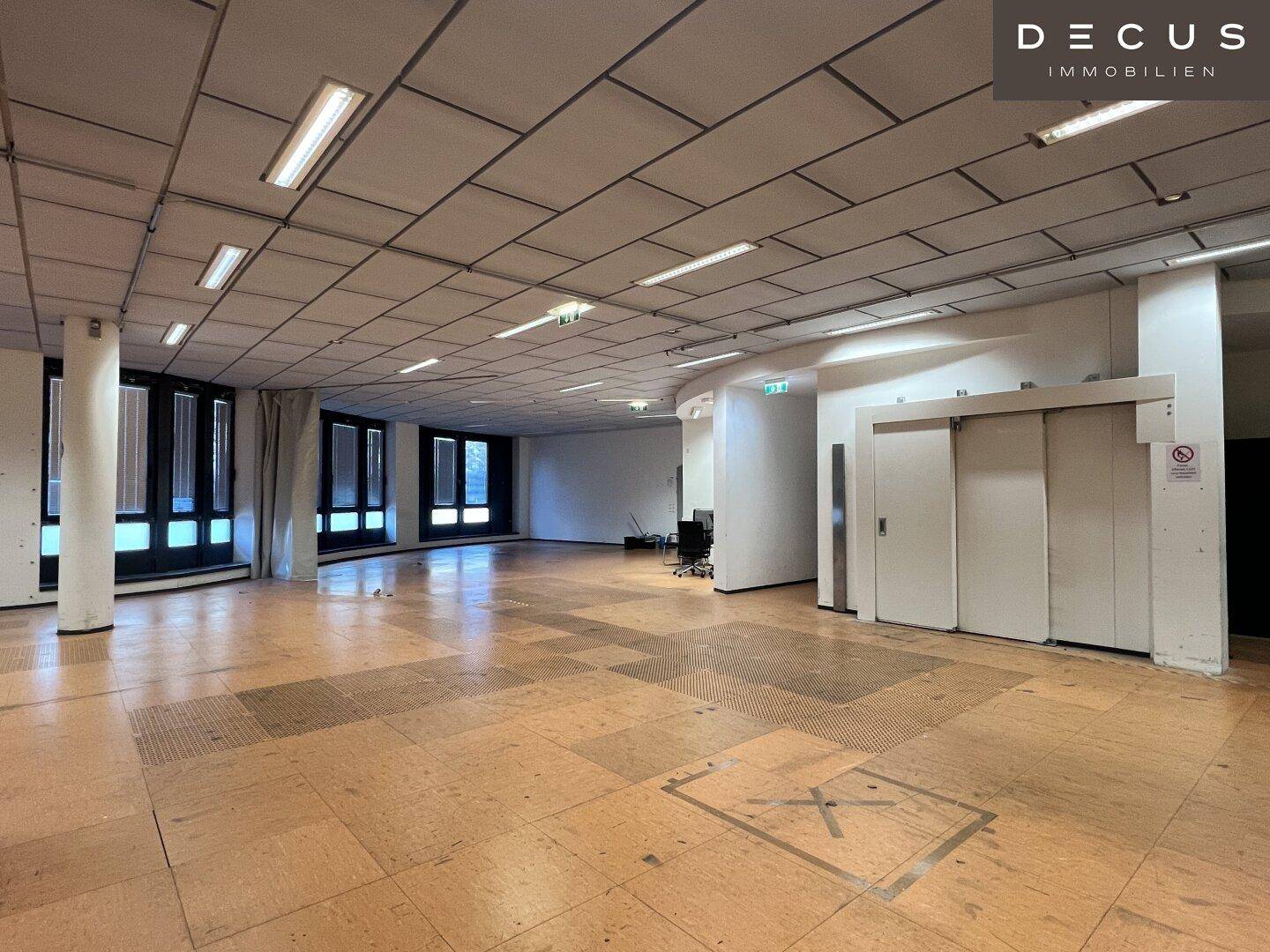 Offener Büroraum mit Säulen, Aufzugstüren und großen Fenstern, die viel Licht hereinlassen.