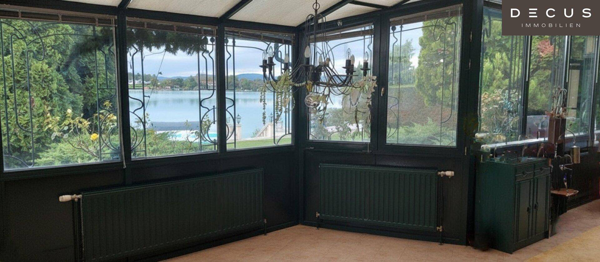 Lichtdurchfluteter Wintergarten mit großen Fenstern, Seeblick und dunklen Heizkörpern.