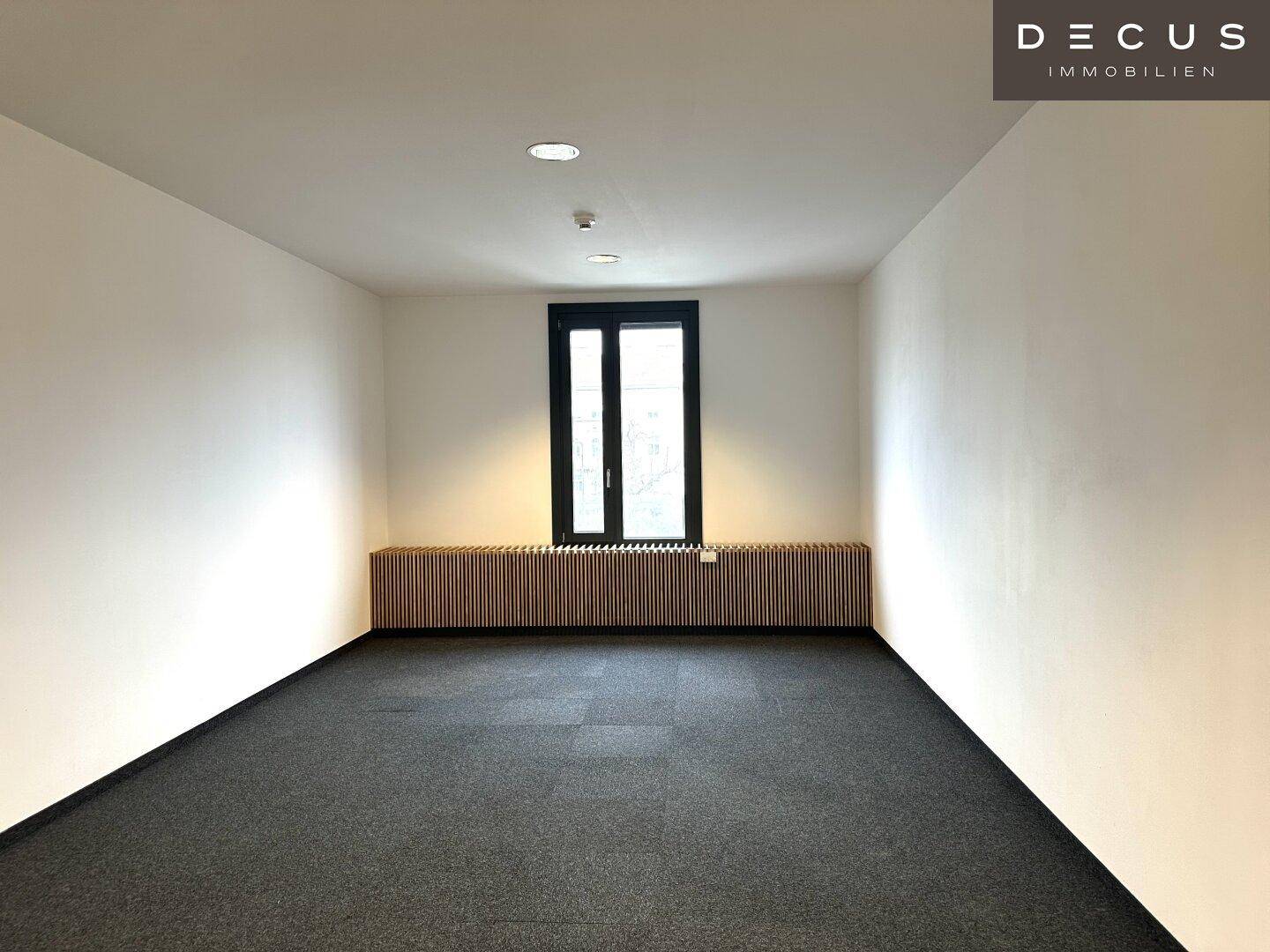 Leerer Büroraum mit Teppichboden, einem Fenster und Holzverkleidung unter dem Fenster.