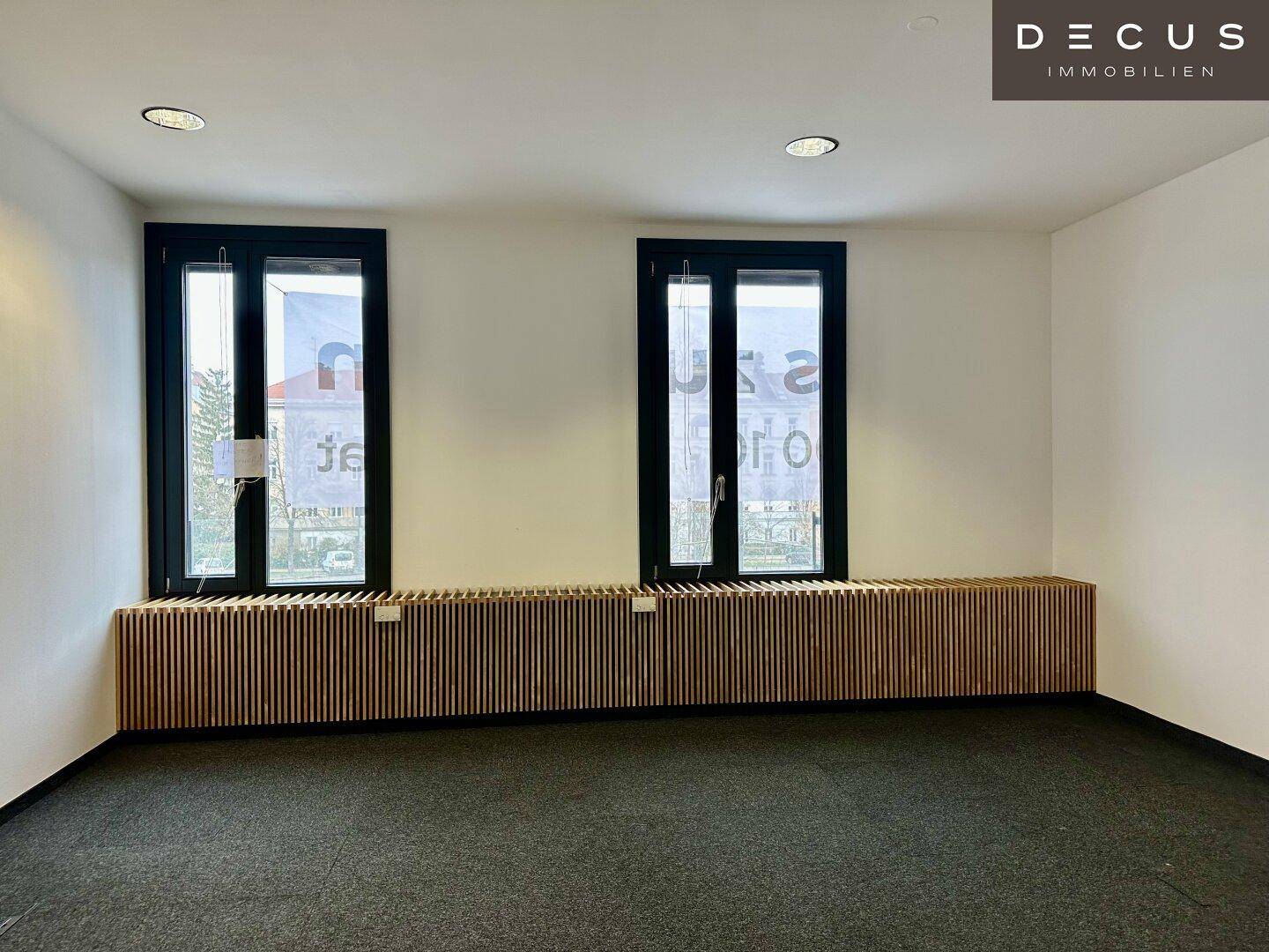 Leerer Büroraum mit zwei Fenstern, Teppichboden und Holzverkleidung unter den Fenstern.