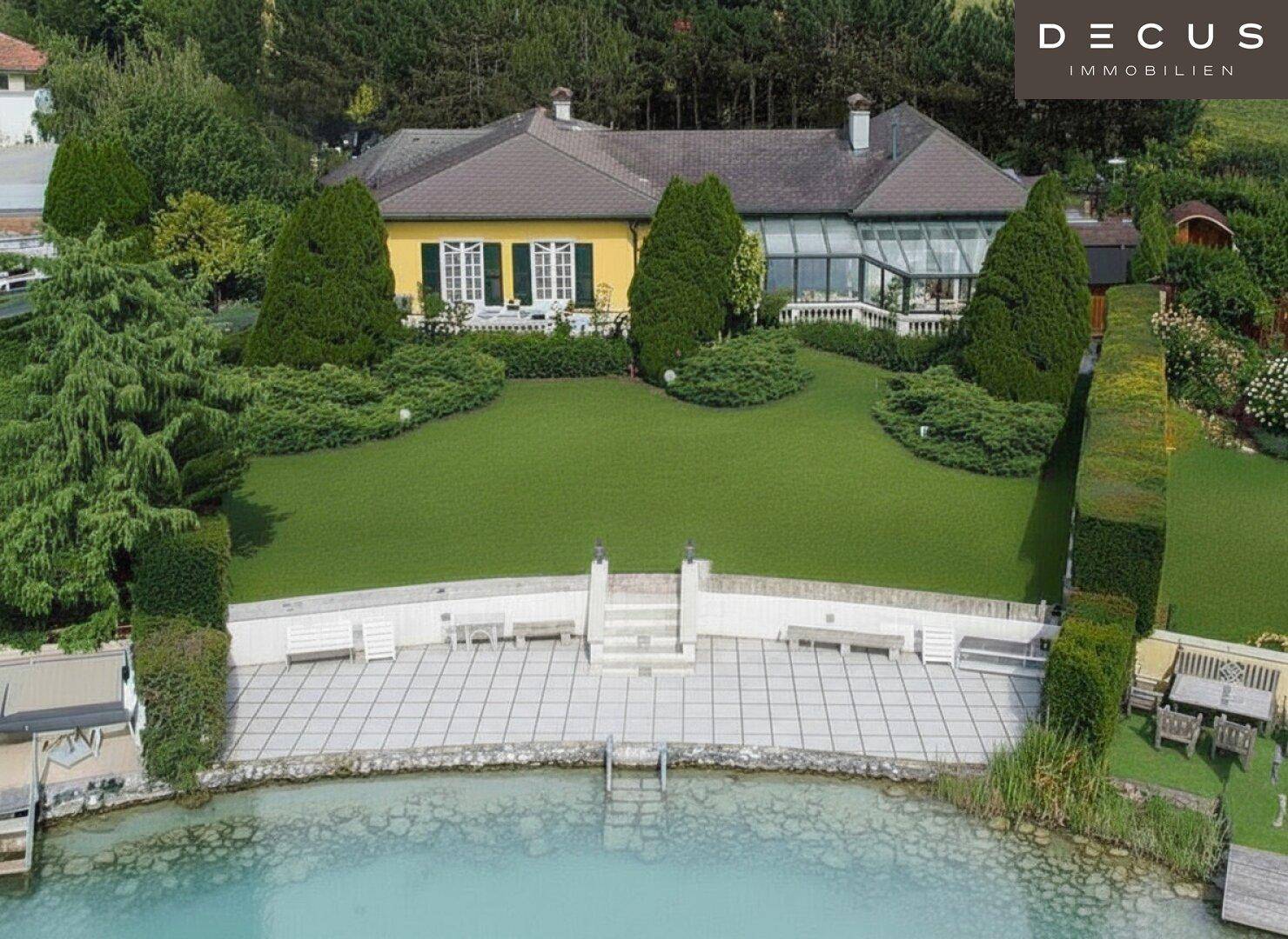 Ansicht des Hauses von der Gartenseite mit großem Rasen, Wintergarten und gepflastertem Uferbereich.