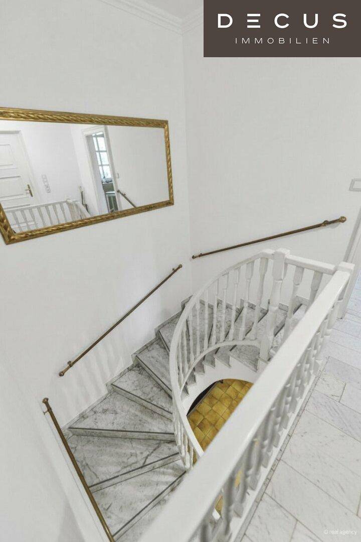 Geschwungene Treppe mit Marmorstufen und weißem Holzgeländer in einem hellen Flur.