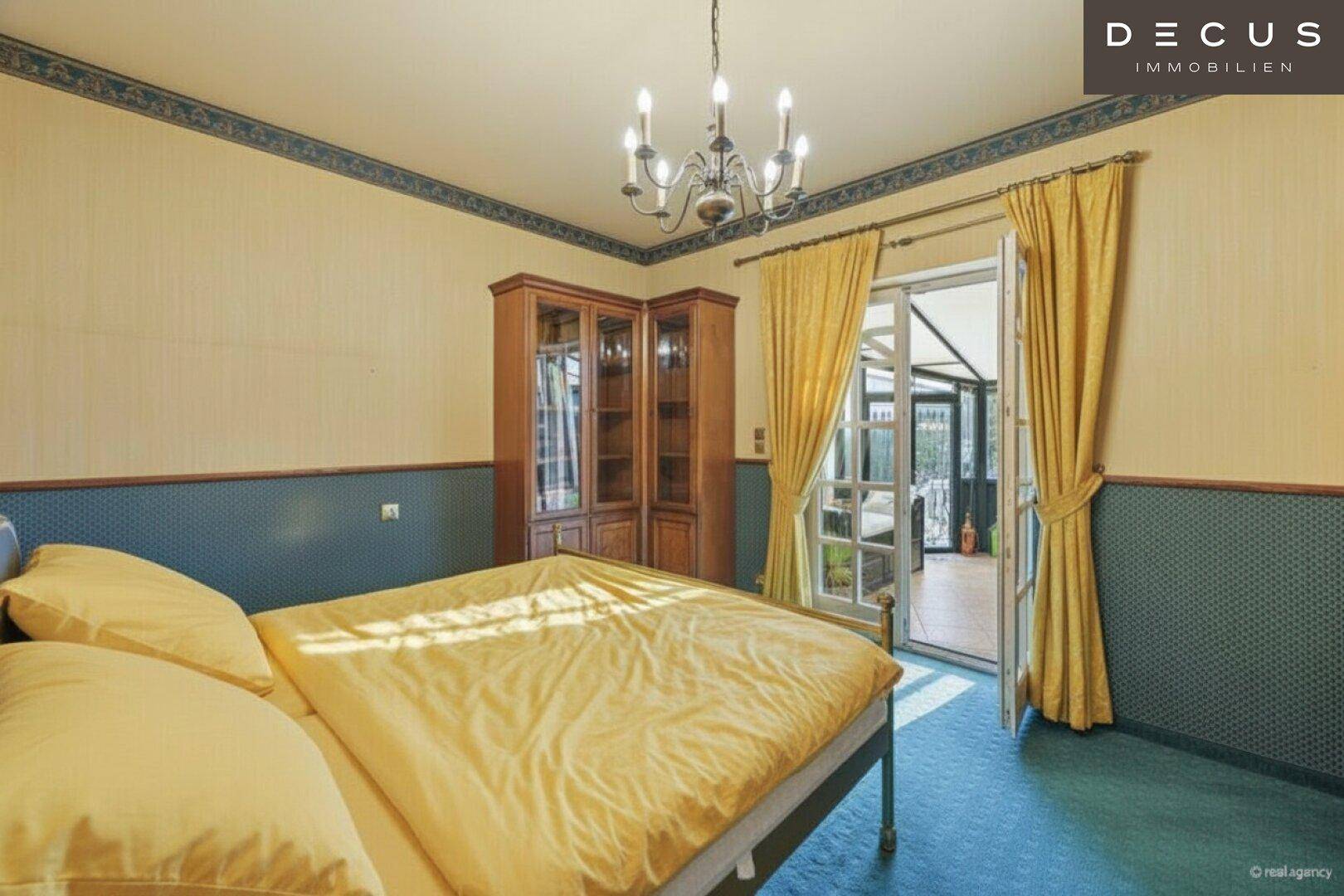 Schlafzimmer mit Bett, Holzschrank und Zugang zum Wintergarten, in blauen und gelben Farbtönen.