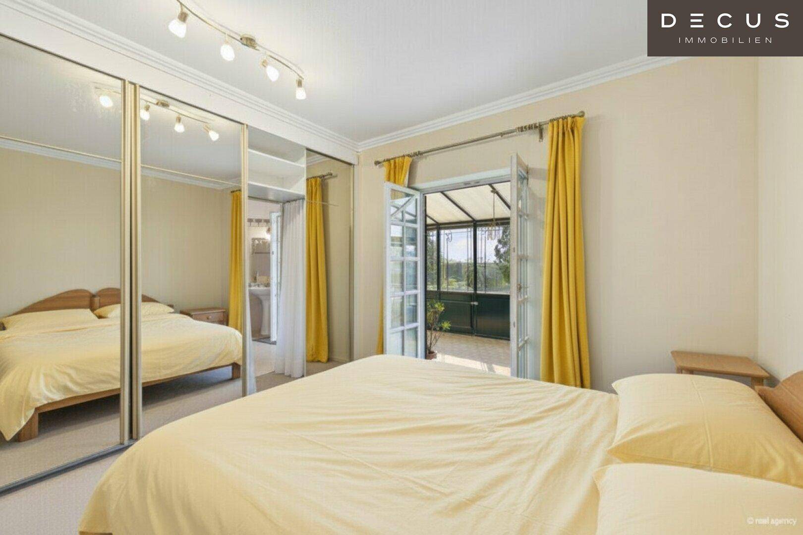 Schlafzimmer mit Bett, Spiegelschränken und Zugang zu einem hellen Wintergarten.