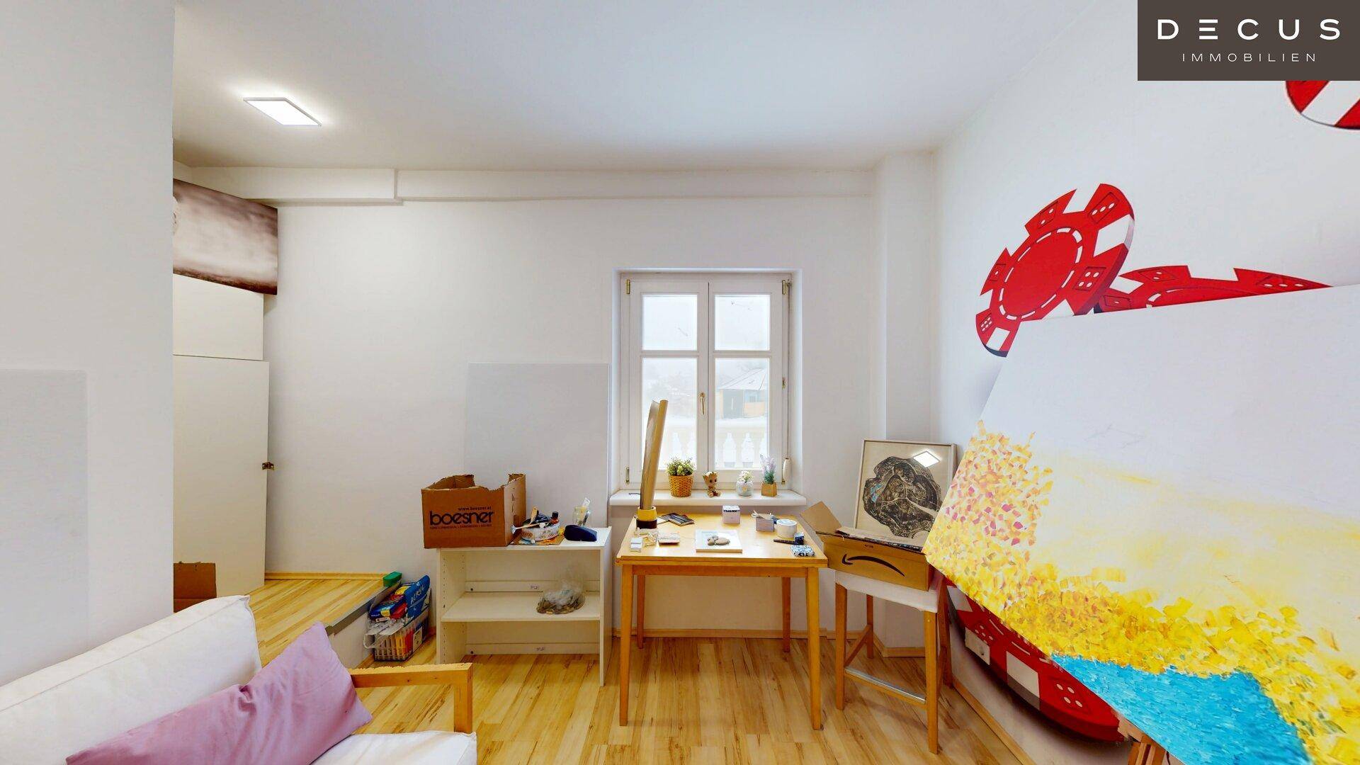 Kreativer Arbeitsbereich mit Schreibtisch, Kunstwerken und viel Tageslicht durch das Fenster.