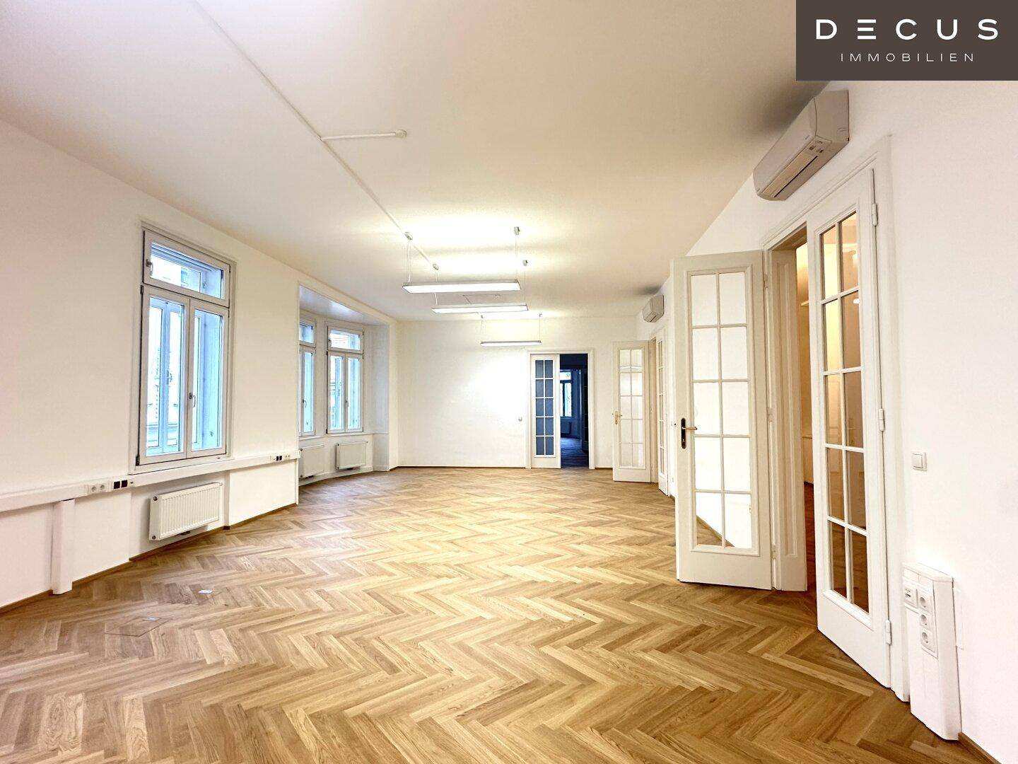 Großer, heller Raum mit Fischgrätparkett, vielen Fenstern und Klimaanlage, ideal für Büronutzung.