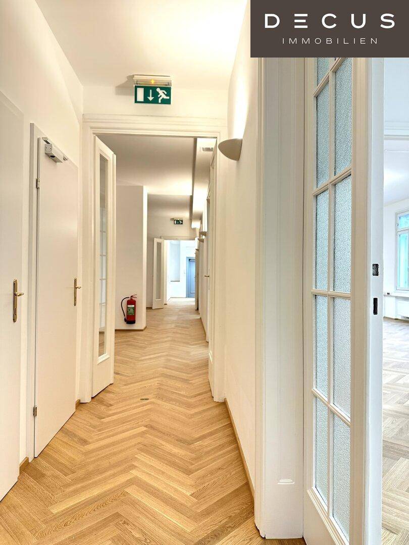 Langer Flur mit Fischgrätparkett, vielen Türen und Notausgangsschild, gut beleuchtet.