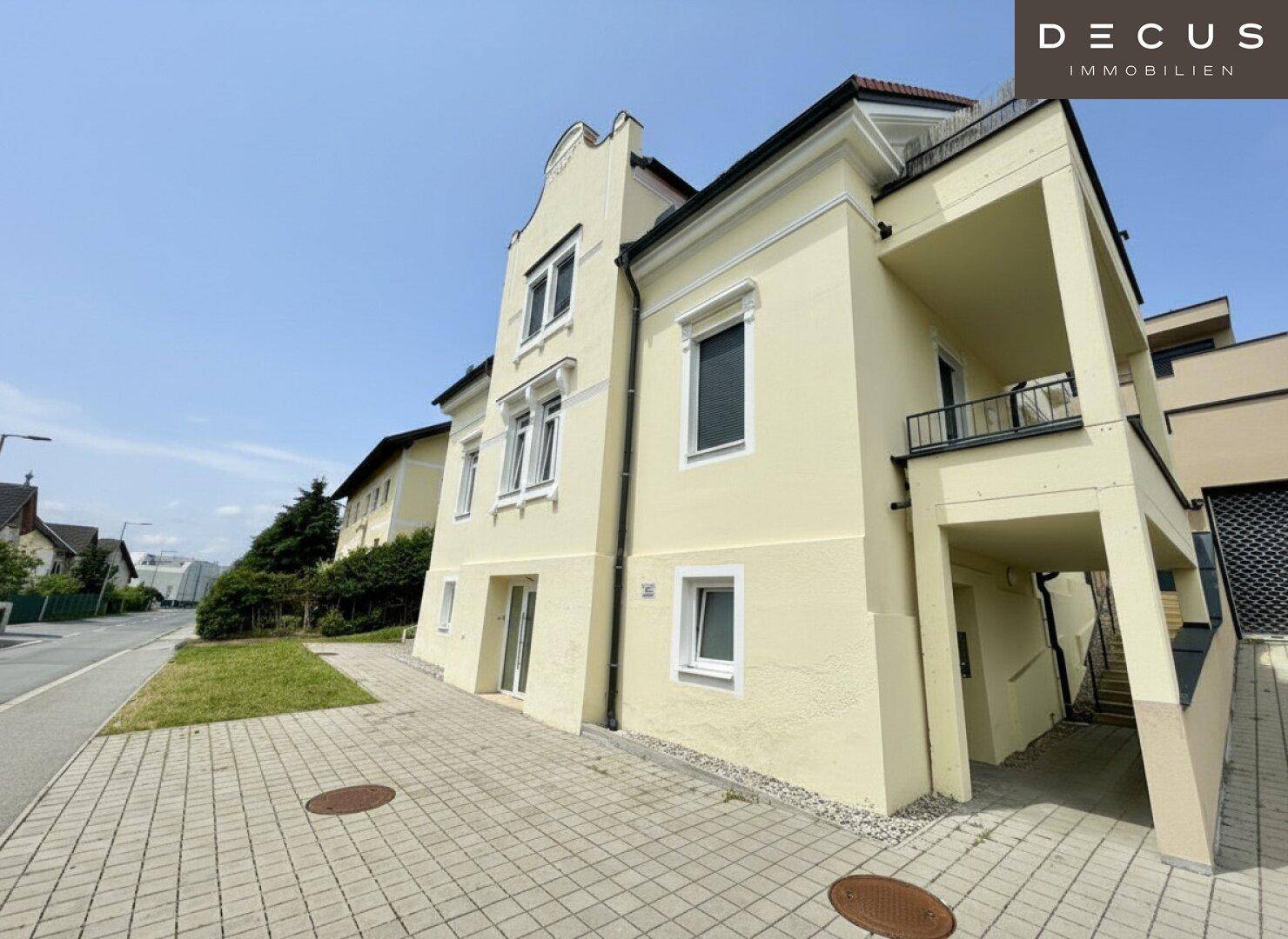Seitenansicht des Gebäudes mit gelber Fassade, gepflastertem Vorplatz und Zufahrt.