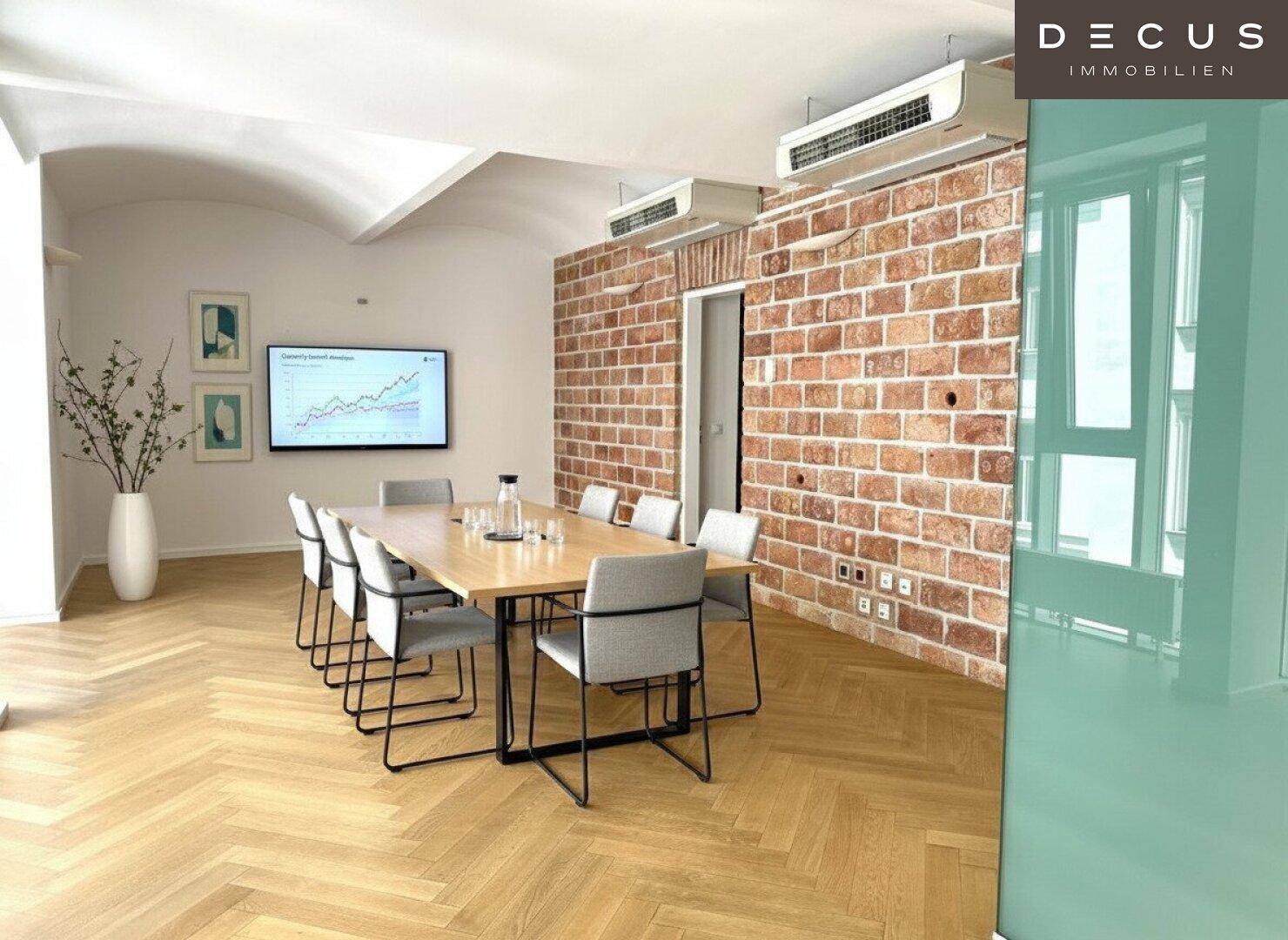 Konferenzraum mit langem Holztisch, Backsteinwand, gewölbter Decke und Fischgrätparkett.