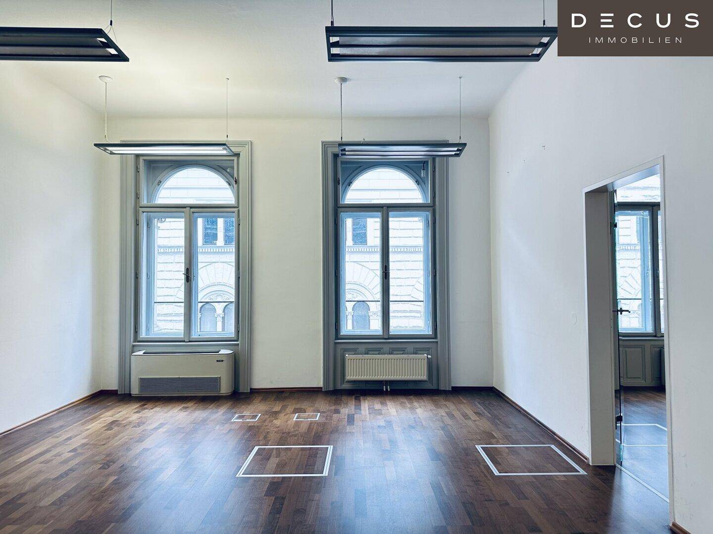 Leeres Büro mit dunklem Holzparkett und hohen Bogenfenstern, bietet viel natürliches Licht.