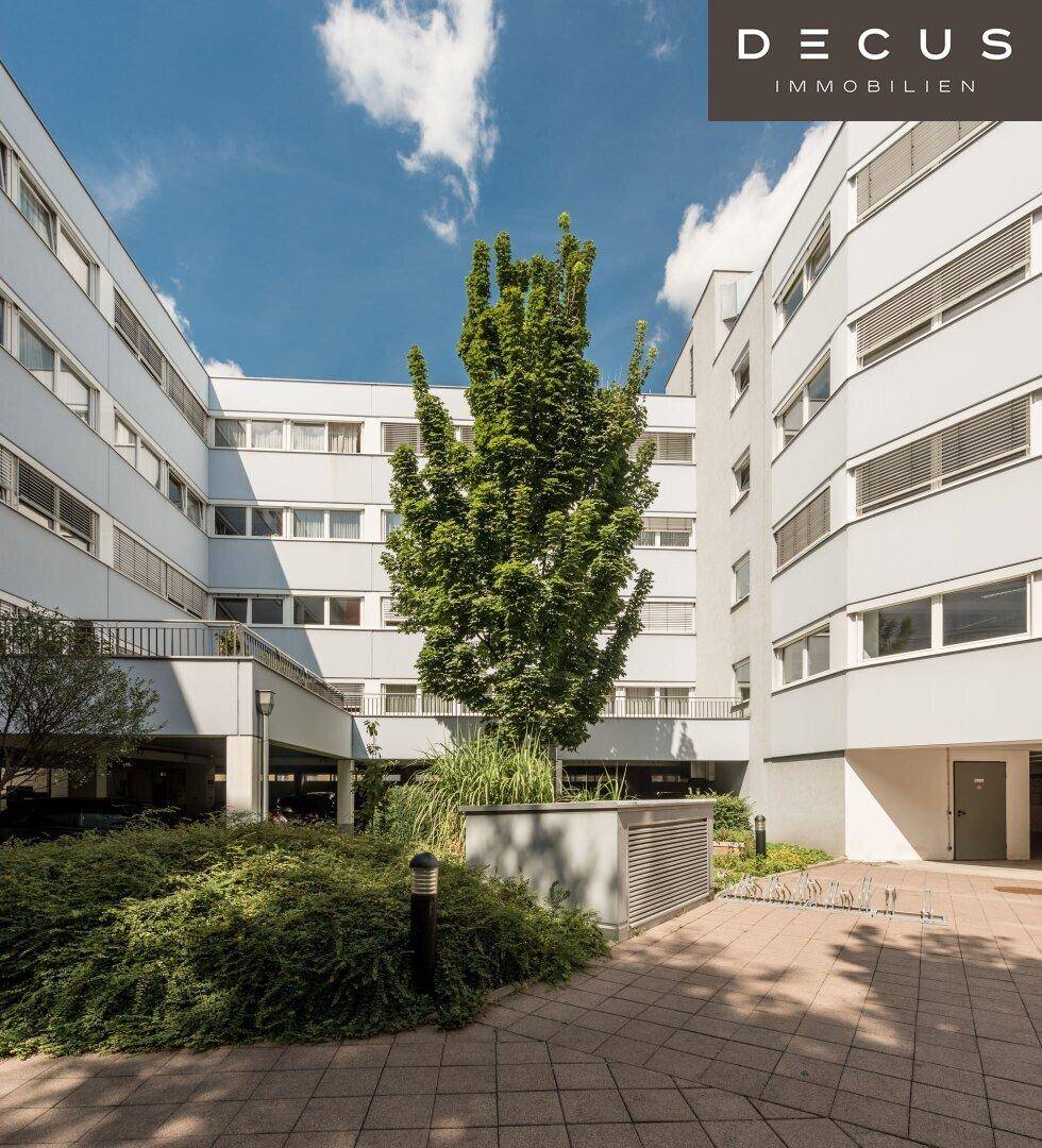Grüner Innenhof eines modernen Gebäudekomplexes mit einem großen Baum und Sträuchern.
